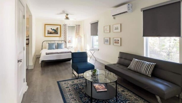 A modern hotel room with a bed, a blue armchair, a black leather sofa, a glass coffee table, and a large window with blinds. - The Blue Heron-elegant urban studio in city center (Memphis)