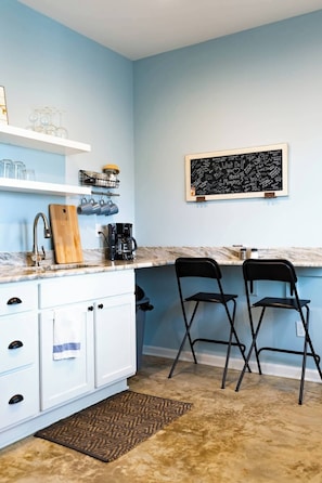 Une cuisine avec un plan de travail en marbre, deux chaises noires, un tableau noir avec des inscriptions et une machine à café.