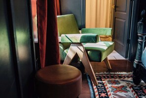 Una habitación con piso de madera, un tapete estampado, una silla verde, un taburete de madera y una puerta con cortina.
