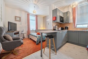 A compact living space with a kitchenette, a bed with a colorful headboard, a gray armchair, a small table, and a bar stool. - First Floor Studio Apartment at Sea-Cote (Blackpool)