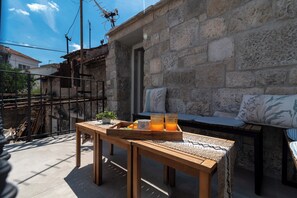 Une table en bois sur laquelle se trouve un plateau avec du jus d’orange et un pot de fleurs, un mur en pierre et un banc avec des coussins.