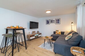 Un salon moderne comprenant un canapé, une table à manger avec des tabourets, un téléviseur fixé au mur et une petite table d’appoint.