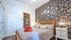 Ein Schlafzimmer mit einem Bett, einem Holzkopfteil, einem Nachttisch mit Lampe, einer Kommode und floraler Tapete.