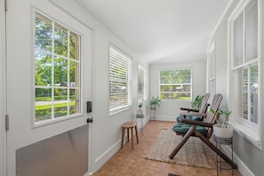 A bright hallway with large windows, a door, a small stool, a chair, and a potted plant.