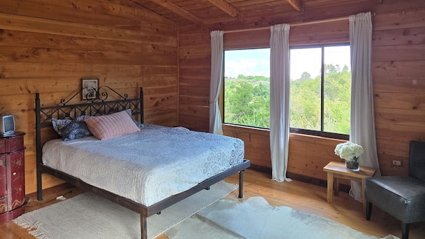 A wooden cabin bedroom with a bed, a chandelier, a window with curtains, a small table with a vase, and a chair. - Panoramic Mountain Views - Luxury Hilltop Retreat in Jardín de Dota (San José)