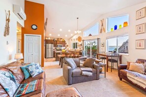 A spacious living area with a sofa, a coffee table, and a dining table. There is a kitchen in the background, and large windows provide natural light.