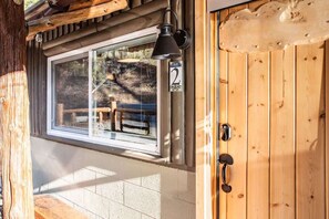 Una puerta de madera con una placa de número, una ventana que da a un banco y árboles, y una lámpara fijada a la pared.