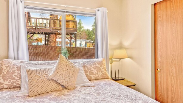 A bed with a patterned bedspread and decorative pillows, a bedside lamp, a window with a view of a wooden deck and trees, and a wooden door. - Walk to Downtown | Family-Friendly Condo | Dogs OK (Leavenworth)