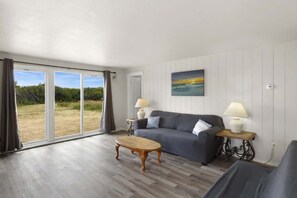 Ein Zimmer mit grauem Sofa, zwei Beistelltischen mit Lampen, einem Holztisch und einem großen Fenster mit Vorhängen.