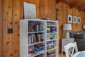 A room with wooden walls, a bookshelf filled with books and board games, a white side table with a lamp, and a grey sofa.