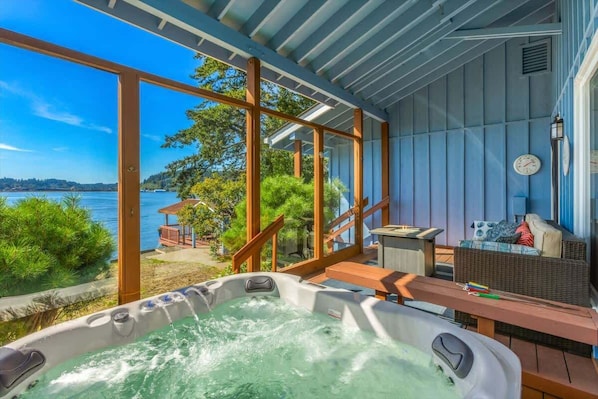 A hot tub with a view of a lake, wooden deck, wicker furniture, and a clock on the wall.