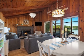A cozy cabin interior with a fireplace, two sofas, a dining table, and large windows offering a view of a lake. - Waterfront Haven: Beach Access, Hot Tub, Fire Pit (Shelton)