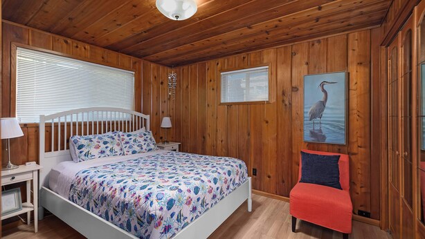 A wooden-paneled room with a bed, bedside tables, a nightstand, a window with blinds, a wall-mounted picture, and a red chair. - Waterfront Haven: Beach Access, Hot Tub, Fire Pit (Shelton)