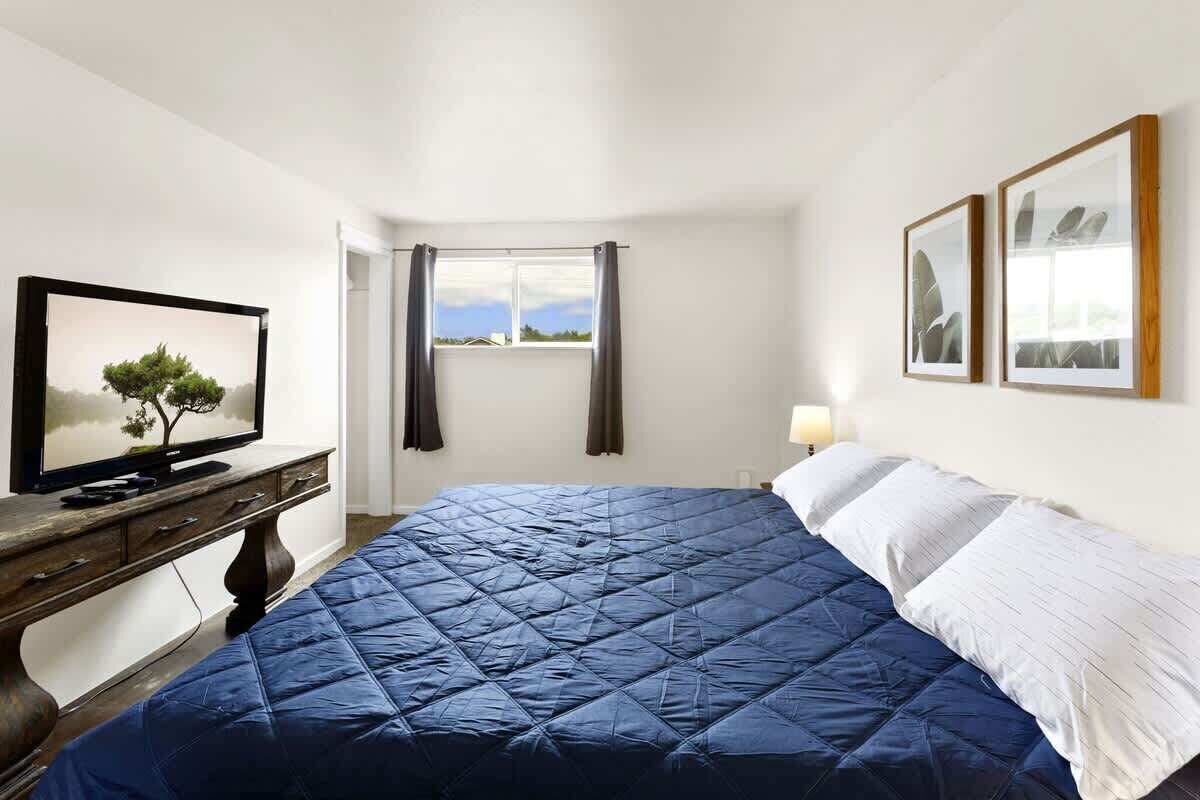 A bedroom with a bed, a television on a wooden dresser, a window with curtains, and two framed pictures on the wall.