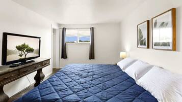 A bedroom with a bed, a television on a wooden dresser, a window with curtains, and two framed pictures on the wall.