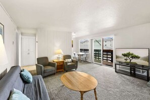 Ein modernes Wohnzimmer mit einer Couch, Sesseln, einem Couchtisch, einem Fernseher und einer Glastür, die auf einen Balkon führt.