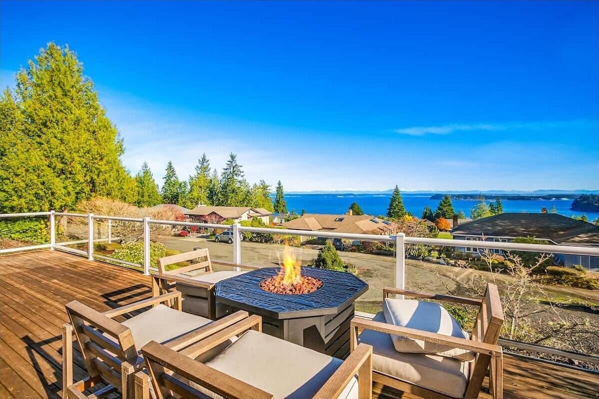 A deck with outdoor furniture, a fire pit, and a view of the ocean and houses.