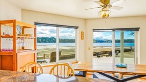 A dining area with a view of the beach, a ceiling fan, and a wooden shelf with glassware.