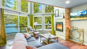 Un salón amplio con un sofá grande, una chimenea y vistas a un lago a través de ventanas enormes.
