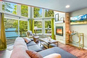 A spacious living room with a large sofa, a fireplace, and a view of a lake through large windows.