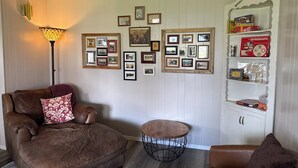 A living room with a brown leather sofa, a small round table, and a wall decorated with framed pictures and a shelf with books and games.