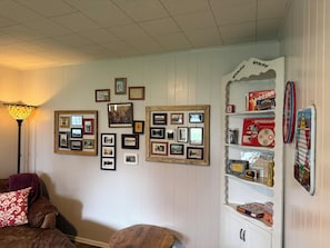 Une pièce dont un mur est décoré de photographies encadrées, avec une lampe de style rétro, un meuble en bois contenant divers objets et un plaid à motifs sur un canapé.