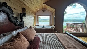 Quarto com cama grande, teto de madeira, vista para um lago e banheira de hidromassagem.