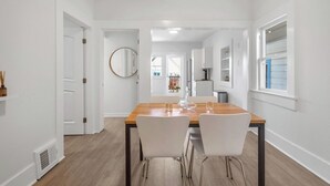 Een moderne eetruimte met een houten tafel en witte stoelen, een ronde spiegel aan de muur en uitzicht op de aangrenzende kamer.