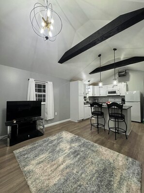 A modern kitchen with white cabinets, a dining area with two bar stools, a flat-screen TV, and a ceiling with exposed beams. - Cozy Tiny Home Unit C (Salem)