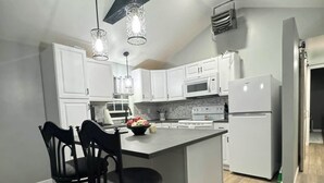 Een moderne keuken met witte kastjes, een centraal eiland, zwarte barstoelen, hanglampen en een zichtbare magnetron.
