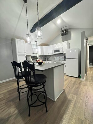 A modern kitchen with white cabinets, a central island, black bar stools, pendant lighting, and a visible microwave. - Cozy Tiny Home Unit C (Salem)
