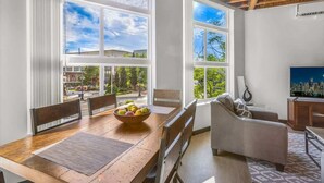 A dining area with a wooden table, chairs, and a bowl of fruit. There are large windows with a view of greenery and buildings. - Bremerton Bay Condo | Walk to Ferry | Unit 201 (Bremerton)