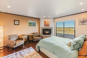 A bedroom with a bed, a fireplace, a desk, two chairs, and a large window with a view.