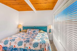 Un dormitorio con cama, techo de madera, paredes blancas y ventana con persianas.