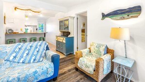 Ein Zimmer mit einem Bett, einem Korbsessel, einem kleinen Tisch, einer Lampe, einer Küchenzeile und einer Wanddekoration mit Fischen.