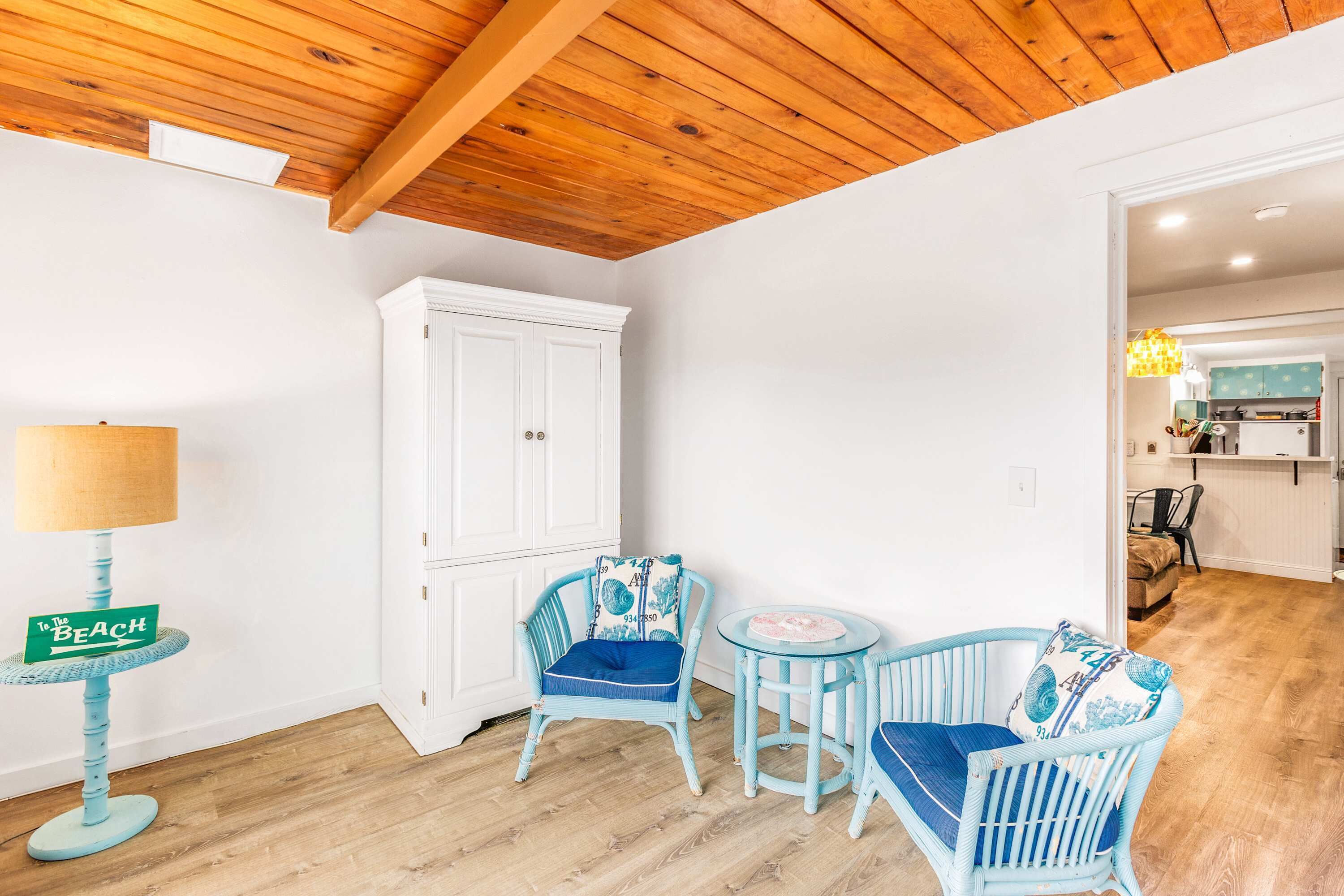 A room with a wooden ceiling, white walls, a white cabinet, two blue chairs with cushions, a small round table, and a lamp with a beach sign.