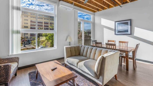 A modern living room with a sofa, dining table, chairs, and large windows offering a view of buildings and trees. - Bremerton Bay Condo | Walk to Ferry | Unit 202 (Bremerton)