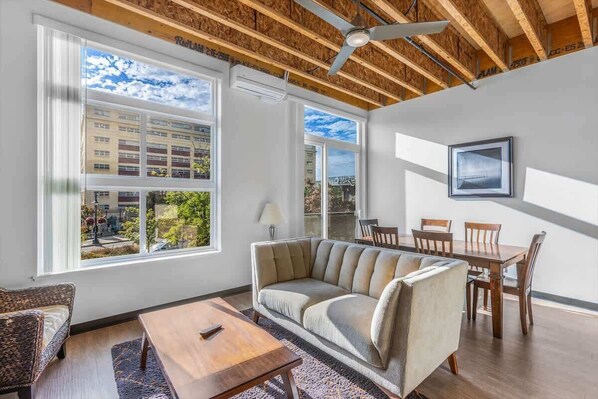 A modern living room with a sofa, dining table, chairs, and large windows offering a view of buildings and trees.