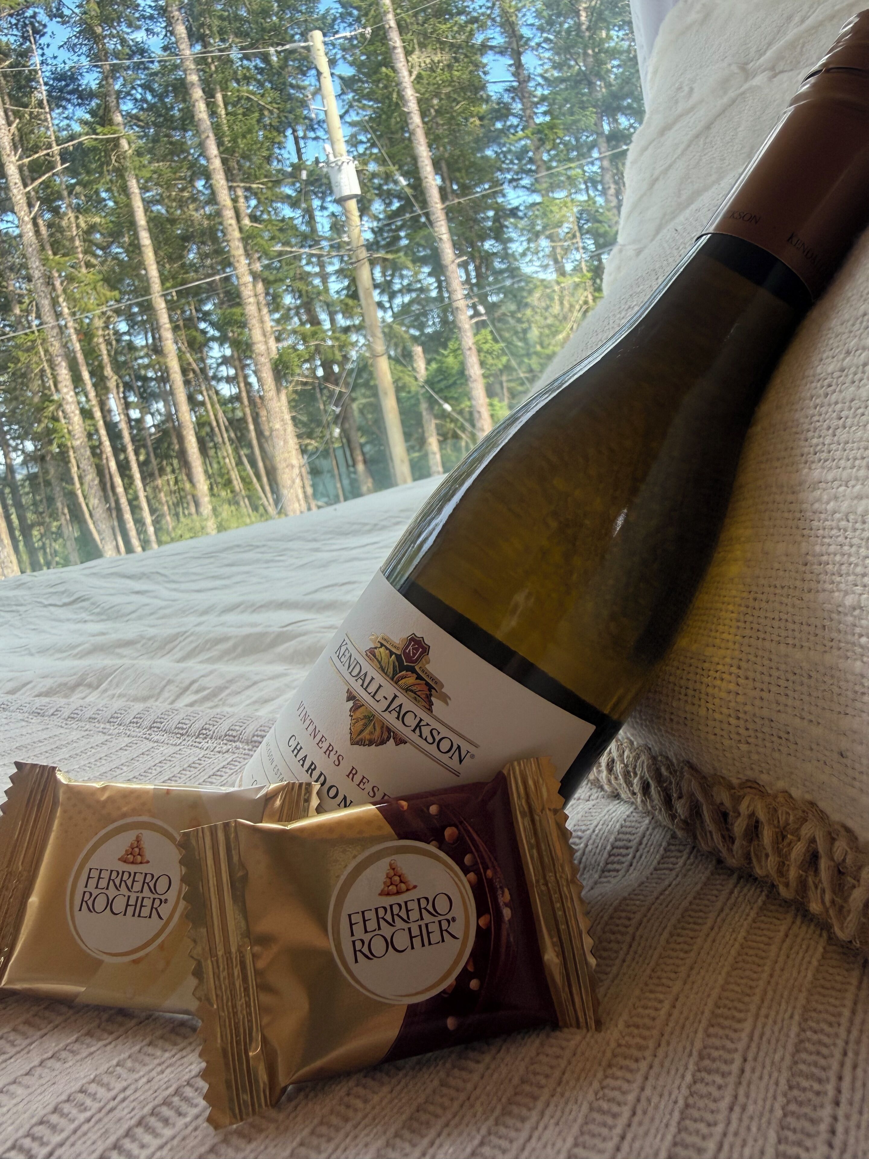 Una ampolla de vi, una etiqueta de vi i dos xocolatines Ferrero Rocher sobre un llit amb vista a un bosc.