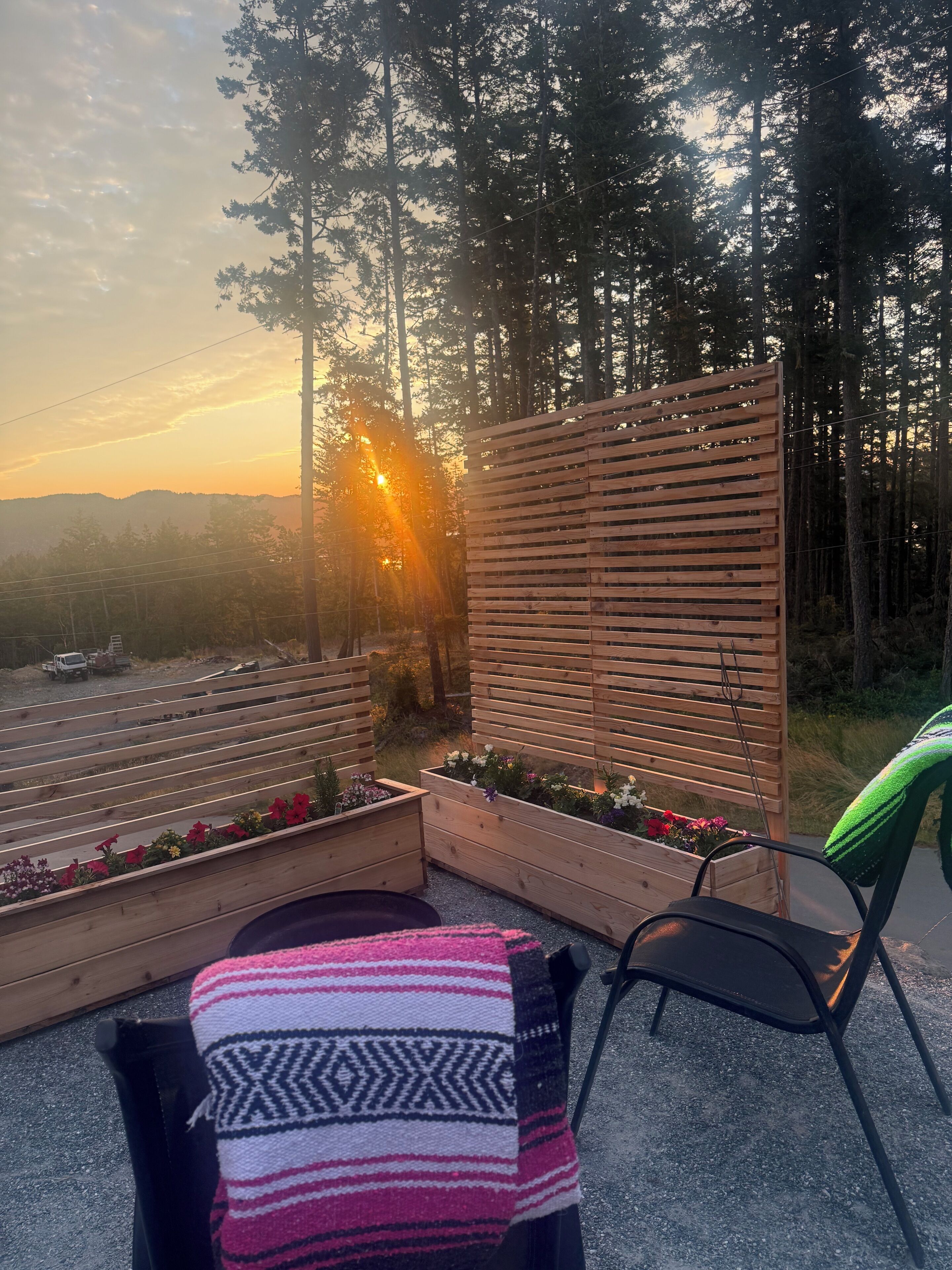 A patio with a view of a forest and a sunset. There are wooden planters with flowers and a black chair.