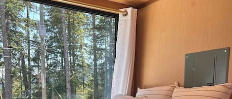 A bedroom with a large window, a bed with multiple pillows, and a view of a forest.