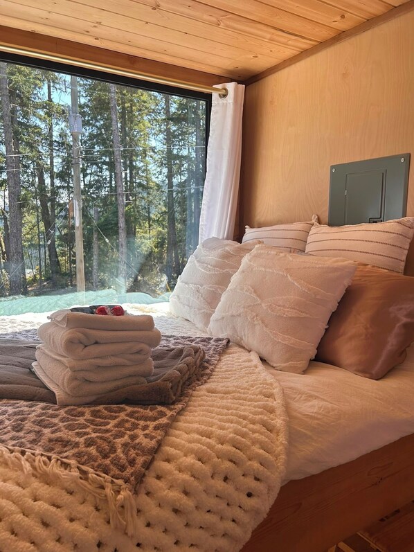 A bedroom with a large window, a bed with multiple pillows, and a view of a forest.
