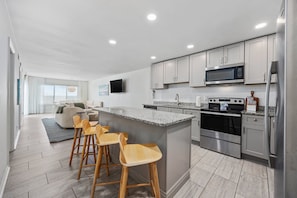 A modern kitchen with a central island, bar stools, stainless steel appliances, and a flat-screen TV on the wall. - The Crestwind - 4BR Oceanfront Unit w/ Pool (Sleeps 10) (North Myrtle Beach)