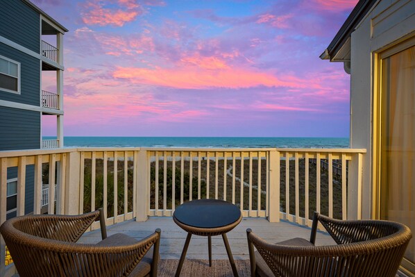 A balcony with wicker chairs and a table, overlooking a beach with a colorful sky. - The Crestwind - 4BR Oceanfront Unit w/ Pool (Sleeps 10) (North Myrtle Beach)