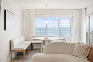 Ein modernes Wohnzimmer mit Sofa, Sitzbank, Esstisch und Meerblick durch große Fenster.