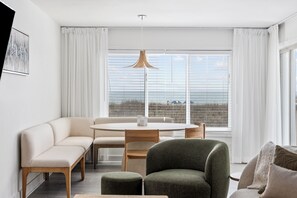 Ein modernes Wohnzimmer mit einer Couch, einem runden Tisch und einem Sessel, einem großen Fenster mit Vorhängen und einer Pendelleuchte.