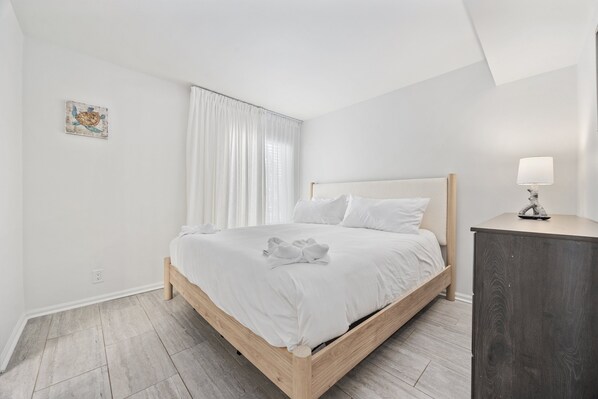 A bedroom with a bed, a bedside table, a lamp, and a wall-mounted artwork. - The Waypoint - 4BR Oceanfront Unit w/ Heated Pool (Sleeps 10) (North Myrtle Beach)