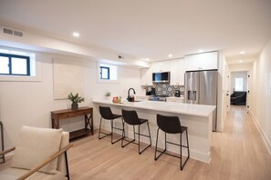 Una cocina moderna con una isla central, taburetes de bar, una mesita de madera y una planta en maceta.