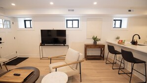 Un salon moderne comprenant un canapé, un téléviseur, une petite table et un coin cuisine avec un évier et des tabourets de bar.