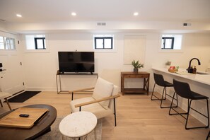 Una sala de estar moderna con un sofá, un televisor, una mesita y una cocina con un fregadero y taburetes de bar.